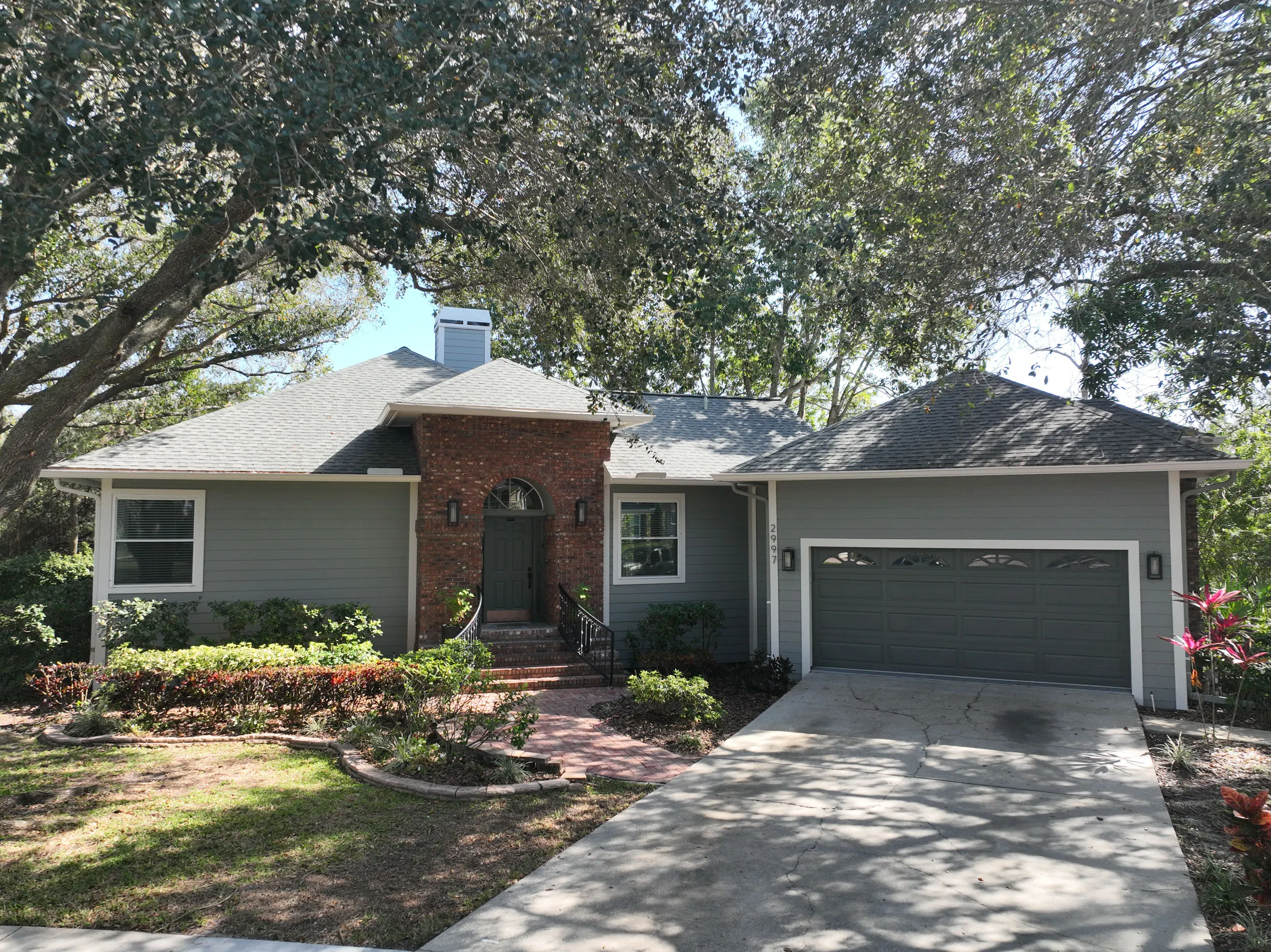 Single-story gray house with a brick front entrance, a two-car garage, and trees casting shadows on the driveway.